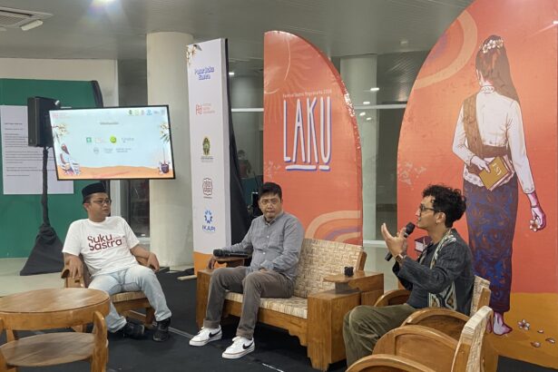 Panel discussion with three men seated on wooden chairs in a bright event space; one speaks into a microphone while another wears a white t-shirt with 'Suku Sastra' on it, backdrop features orange banners with artwork and logos.