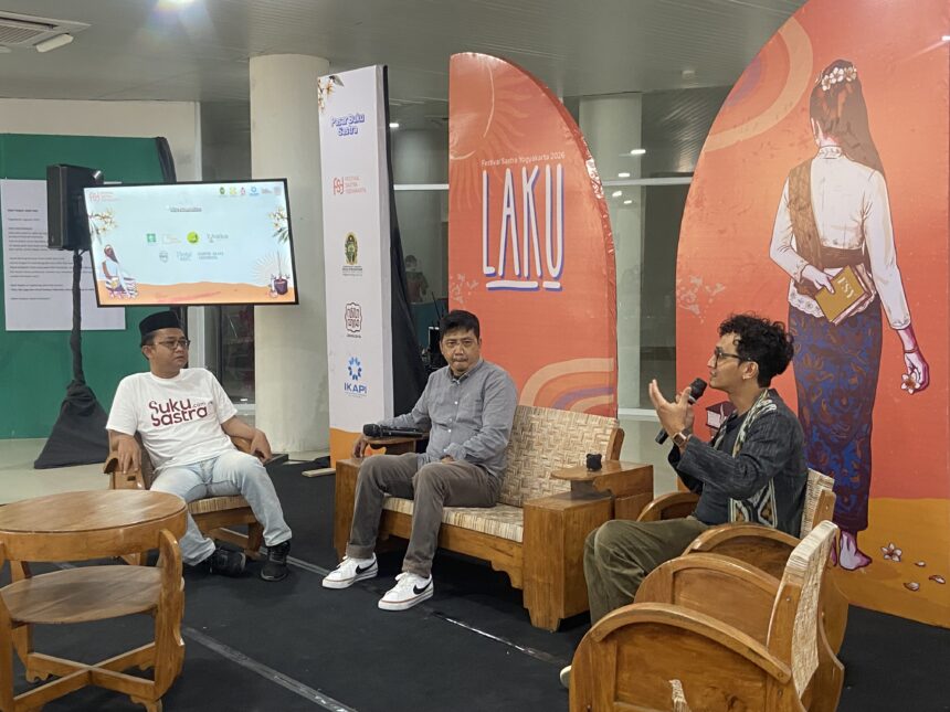 Panel discussion with three men seated on wooden chairs in a bright event space; one speaks into a microphone while another wears a white t-shirt with 'Suku Sastra' on it, backdrop features orange banners with artwork and logos.