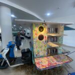Colorful wooden bookshelf sculpture with speakers inside a bright interior; a woman in a hijab stands nearby, blue scooter in foreground.