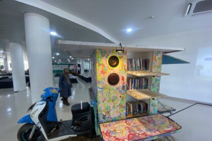 Colorful wooden bookshelf sculpture with speakers inside a bright interior; a woman in a hijab stands nearby, blue scooter in foreground.