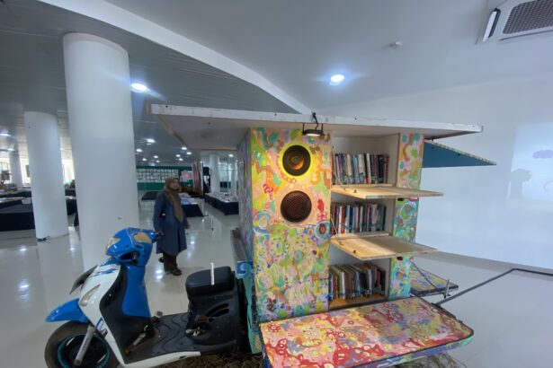 Colorful wooden bookshelf sculpture with speakers inside a bright interior; a woman in a hijab stands nearby, blue scooter in foreground.