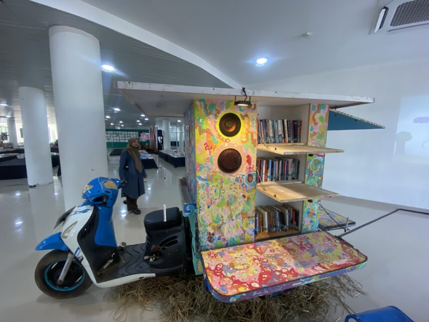 Colorful wooden bookshelf sculpture with speakers inside a bright interior; a woman in a hijab stands nearby, blue scooter in foreground.