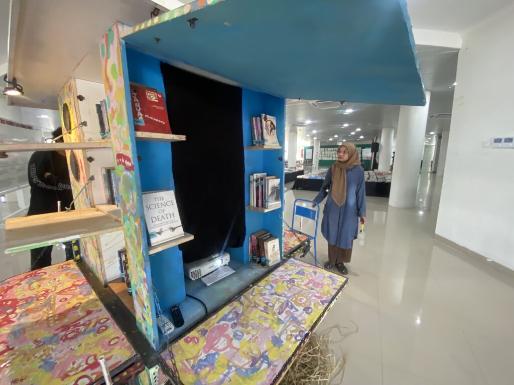 Woman wearing a hijab browses books at a colorful bookstore kiosk in a bright mall setting.
