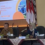 Panel of officials seated at a long table with microphones and nameplates during a formal meeting; presentation screen in the background.
