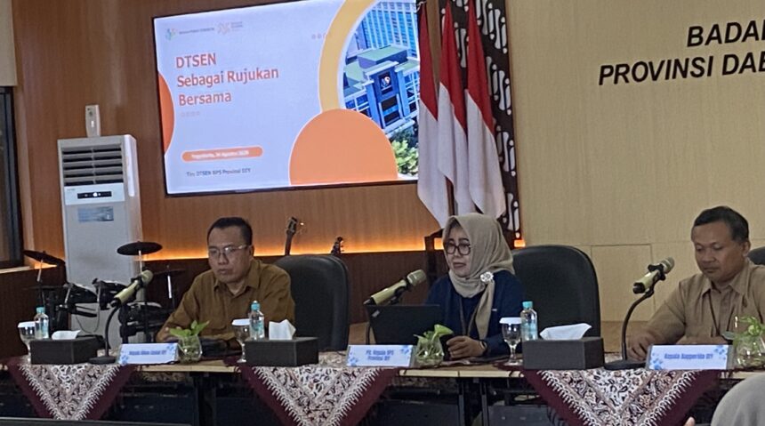 Panel of officials seated at a long table with microphones and nameplates during a formal meeting; presentation screen in the background.