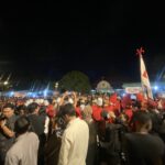 Crowd of people at a night celebration, many wearing red and white, waving flags under bright lights.