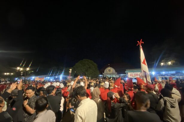 Crowd of people at a night celebration, many wearing red and white, waving flags under bright lights.