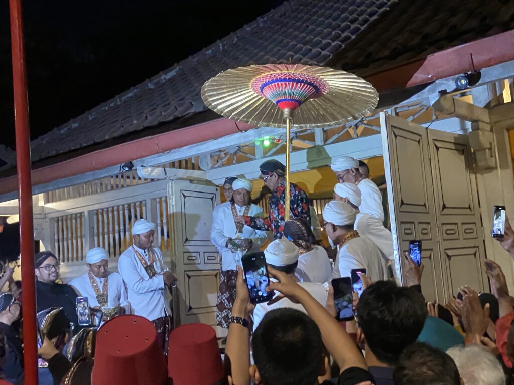 IMG_2620 - Mabur.co Crowd watches a nighttime cultural performance on a stage, with performers in white outfits and turbans under a large decorative parasol or umbrella.