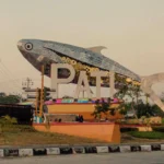 Giant mosaic fish sculpture over the letters PAT at a roundabout, with a bus and road nearby.