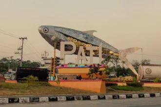 Giant mosaic fish sculpture over the letters PAT at a roundabout, with a bus and road nearby.