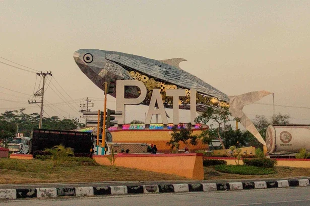 Giant mosaic fish sculpture over the letters PAT at a roundabout, with a bus and road nearby.