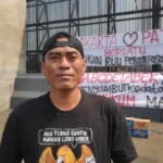 Man in a black cap and black T-shirt stands outdoors in front of a metal fence with large protest banners featuring red and black text in Indonesian; cardboard boxes sit on the ground nearby.