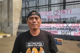 Man in a black cap and black T-shirt stands outdoors in front of a metal fence with large protest banners featuring red and black text in Indonesian; cardboard boxes sit on the ground nearby.