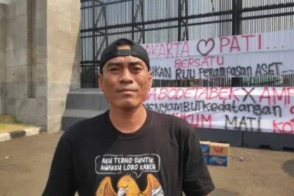 Man in a black cap and black T-shirt stands outdoors in front of a metal fence with large protest banners featuring red and black text in Indonesian; cardboard boxes sit on the ground nearby.
