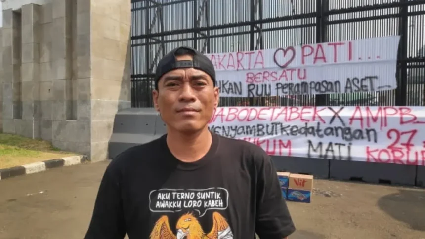 Man in a black cap and black T-shirt stands outdoors in front of a metal fence with large protest banners featuring red and black text in Indonesian; cardboard boxes sit on the ground nearby.