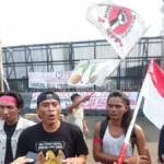 Protesters gather outside a gated area, waving an Indonesian flag and holding banners with Indonesian text.