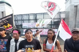 Protesters gather outside a gated area, waving an Indonesian flag and holding banners with Indonesian text.