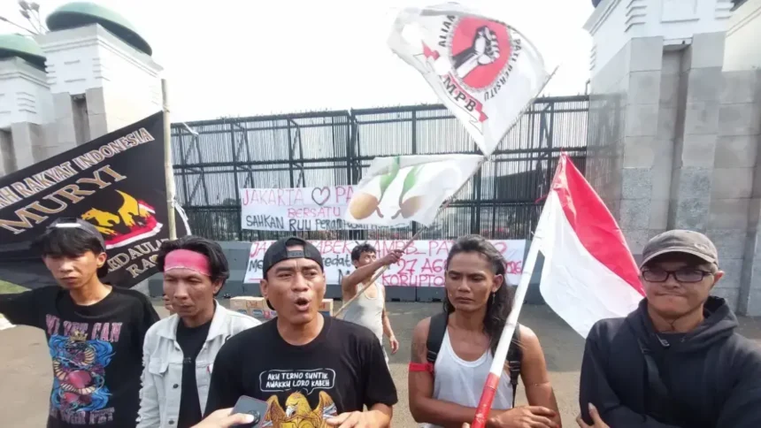 Protesters gather outside a gated area, waving an Indonesian flag and holding banners with Indonesian text.
