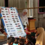 Two women in hijabs explain a large fish species chart to a group of listening children outdoors.