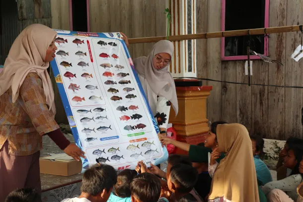 IMG_2774 - Mabur.co Two women in hijabs explain a large fish species chart to a group of listening children outdoors.