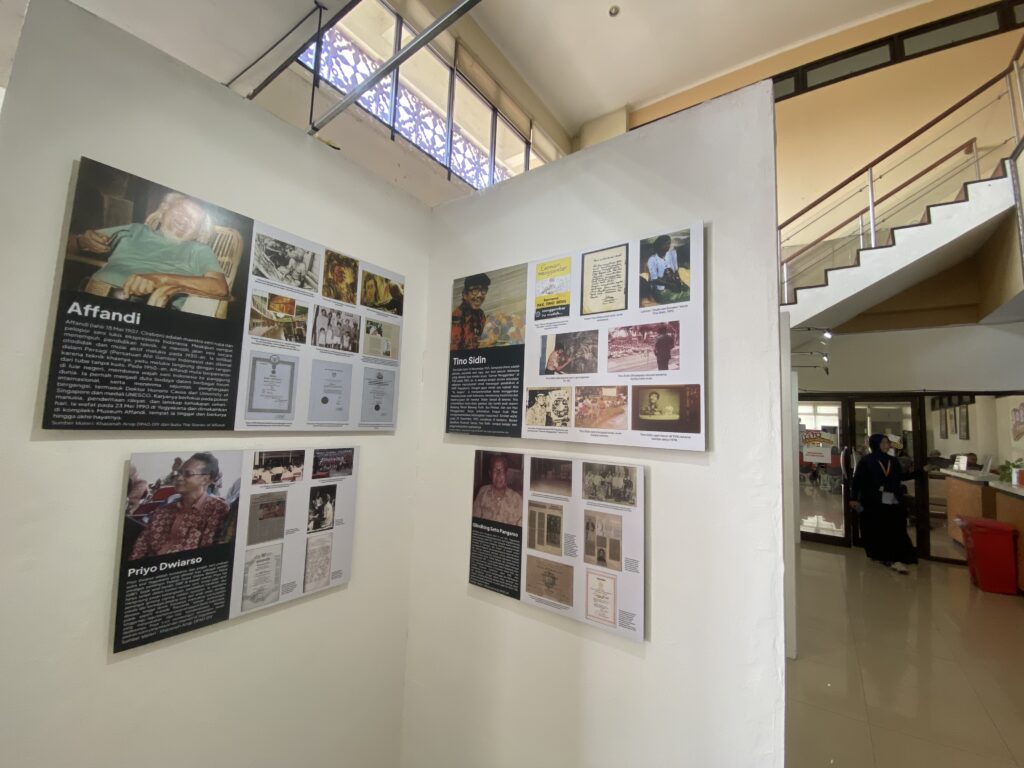 Exhibition hallway with white walls displaying large poster boards about people; a corner wall lines with photos and text, near a staircase and glass railing.