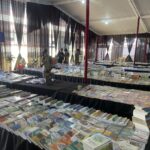 Book fair booth with long tables of magazines and books under a white tent with black sheer curtains; visitors browse.
