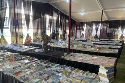 Book fair booth with long tables of magazines and books under a white tent with black sheer curtains; visitors browse.