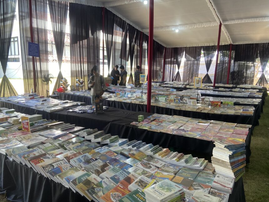 Book fair booth with long tables of magazines and books under a white tent with black sheer curtains; visitors browse.