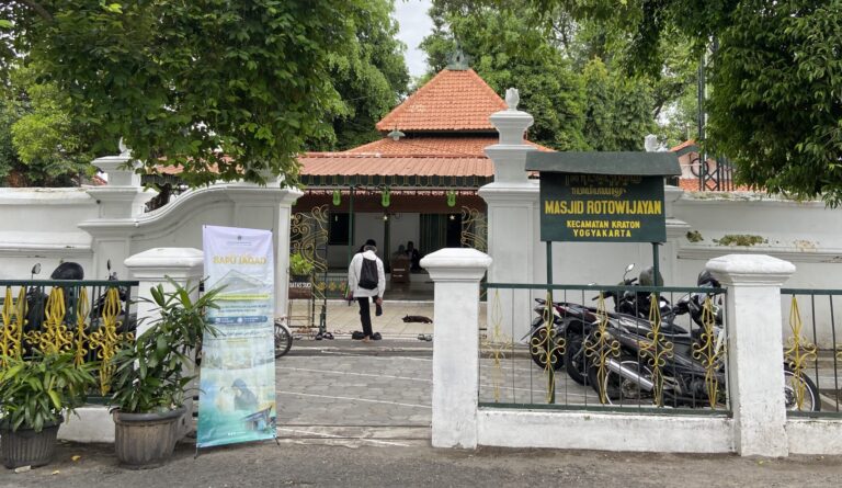 Entrance of Masjid Rotowijayan Kraton Yogyakarta: white walls, red-tiled roof, decorative gate, and a sign near parked motorcycles under leafy trees.