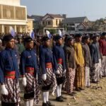 A large group of people standing in rows outdoors in traditional batik outfits with colored sashes and feathered headpieces, likely part of a ceremony or parade in a town square.