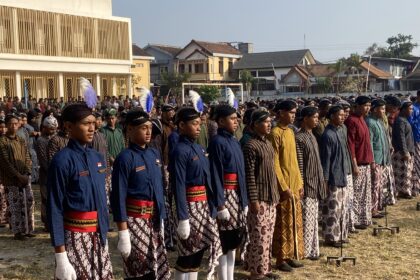 A large group of people standing in rows outdoors in traditional batik outfits with colored sashes and feathered headpieces, likely part of a ceremony or parade in a town square.