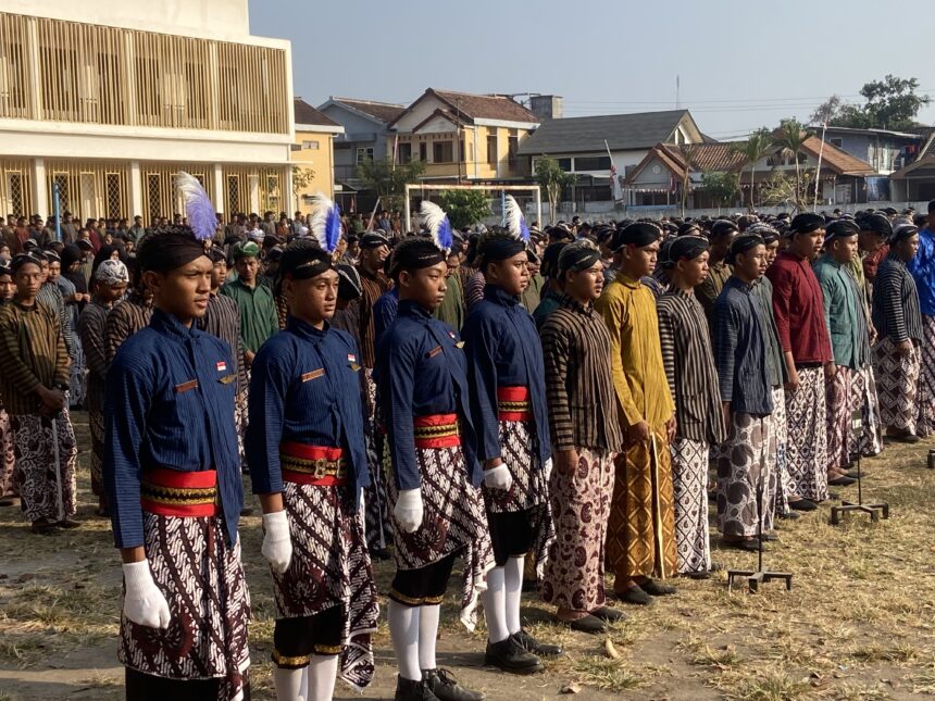 A large group of people standing in rows outdoors in traditional batik outfits with colored sashes and feathered headpieces, likely part of a ceremony or parade in a town square.