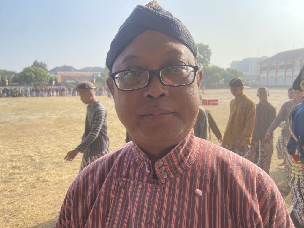 Close-up selfie of a person wearing a dark headwrap and glasses outdoors, with a group and buildings in the background.