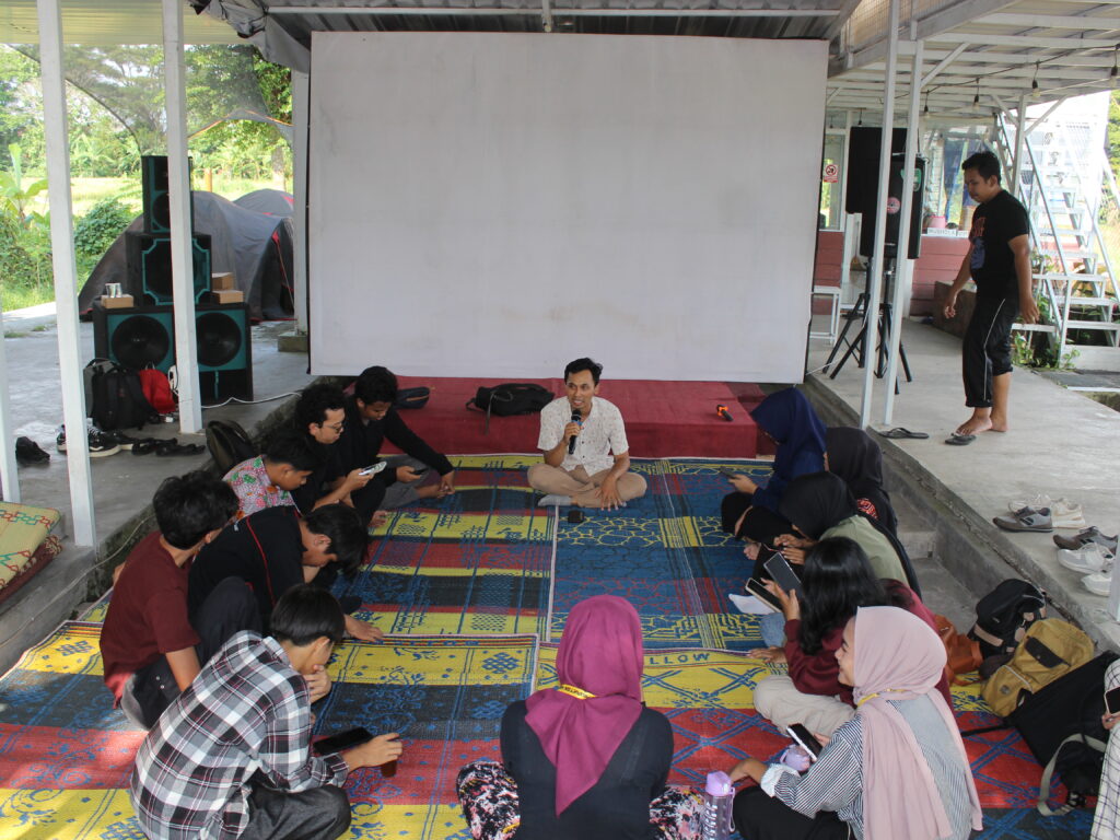 Group of people sitting in a circle on colorful mats under a shelter, a man speaks into a microphone at the center.