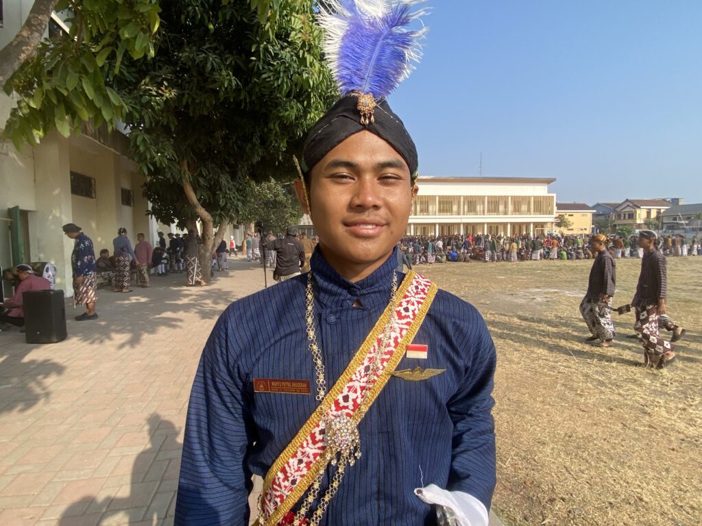 Young man in ceremonial attire with a blue striped outfit and a purple feather headdress at a sunny outdoor festival.