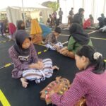 Group of children sitting on a black floor inside a tent, wearing striped outfits and hijabs, working with colorful candies on wooden trays as part of a craft activity