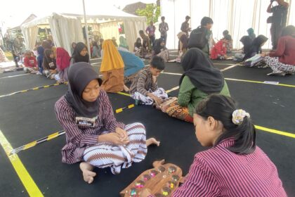 Group of children sitting on a black floor inside a tent, wearing striped outfits and hijabs, working with colorful candies on wooden trays as part of a craft activity