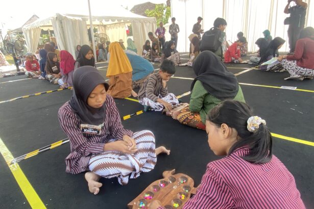 Group of children sitting on a black floor inside a tent, wearing striped outfits and hijabs, working with colorful candies on wooden trays as part of a craft activity