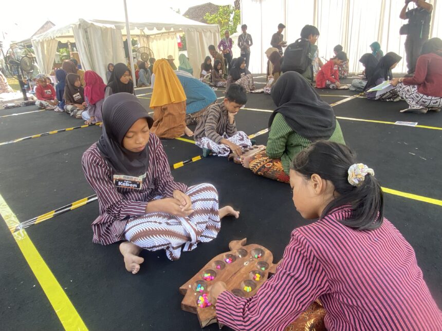 Group of children sitting on a black floor inside a tent, wearing striped outfits and hijabs, working with colorful candies on wooden trays as part of a craft activity