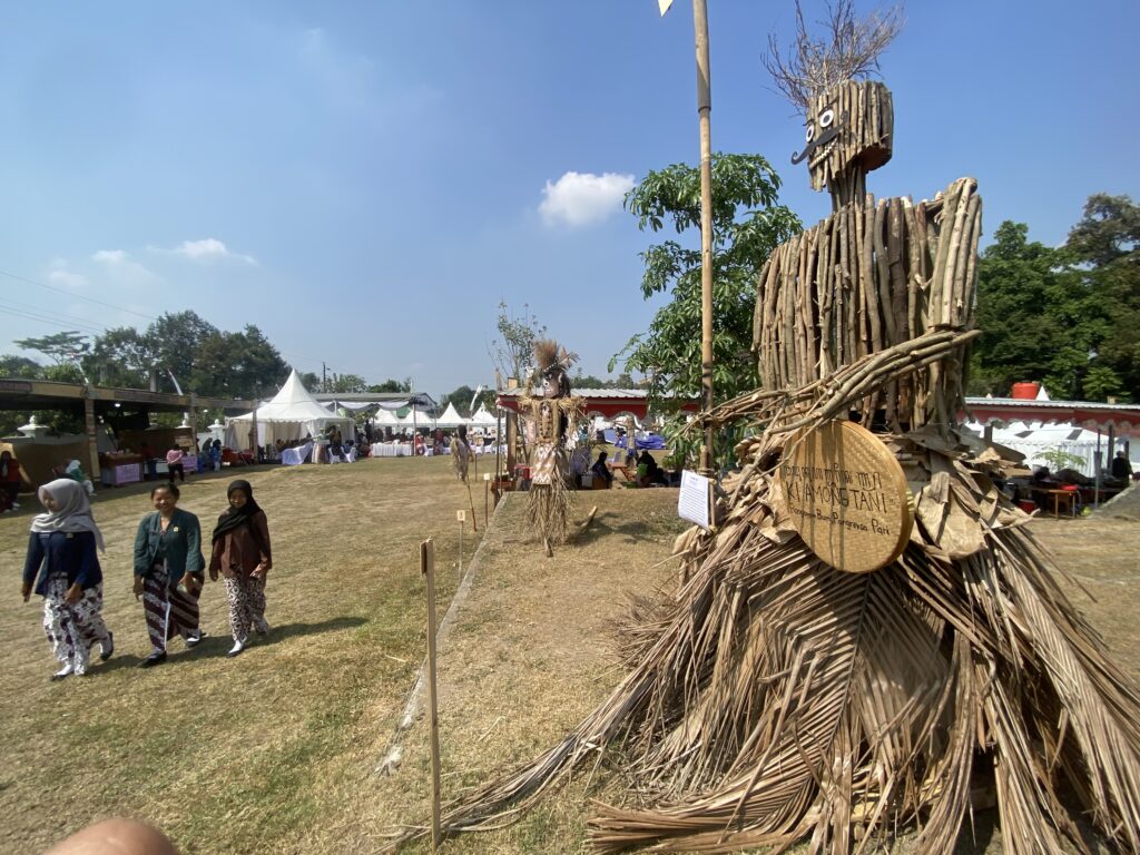 Three women in matching batik dresses walk through a sunny outdoor festival with white tents; a tall straw sculpture stands on the right.