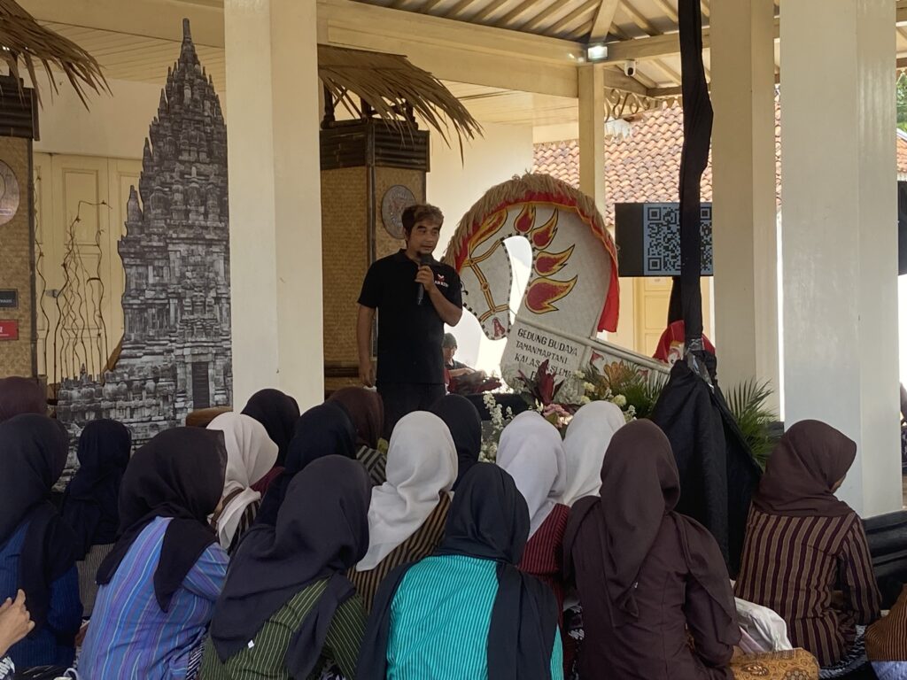 Speaker holding microphone addresses a seated group of students wearing hijabs in a hall with decorative pillars and a large drum display behind him