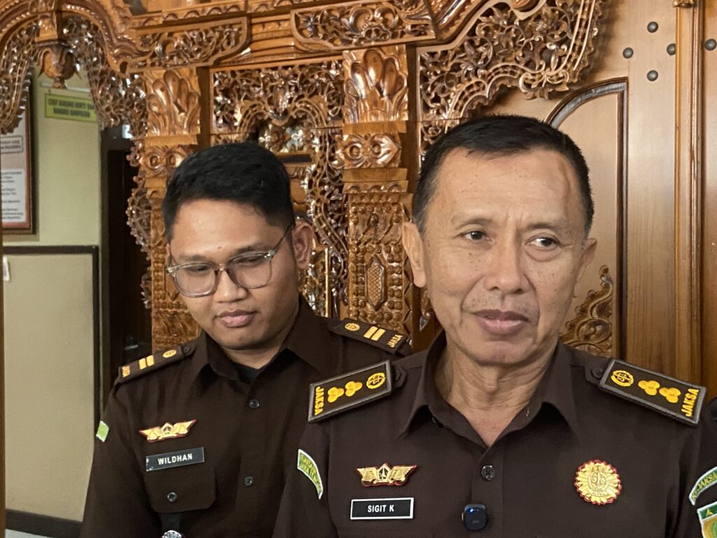 Two uniformed men pose for a photo in front of an intricately carved wooden backdrop, with mission badges on their chests and name tags visible (e.g., Sigit K).