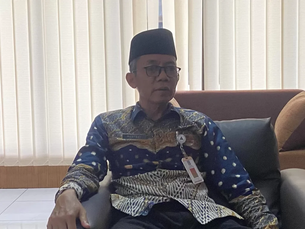 Older man wearing a black cap (peci) and patterned batik shirt sits on a sofa in an office lounge with a name badge clipped to his chest.
