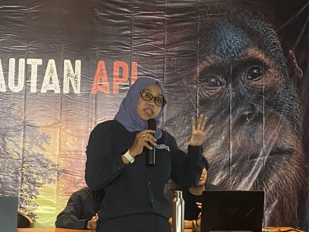 IMG_3093 - Mabur.co A woman wearing a blue hijab speaks into a microphone on a stage with a large banner showing an orangutan image and white/orange letters in the background.