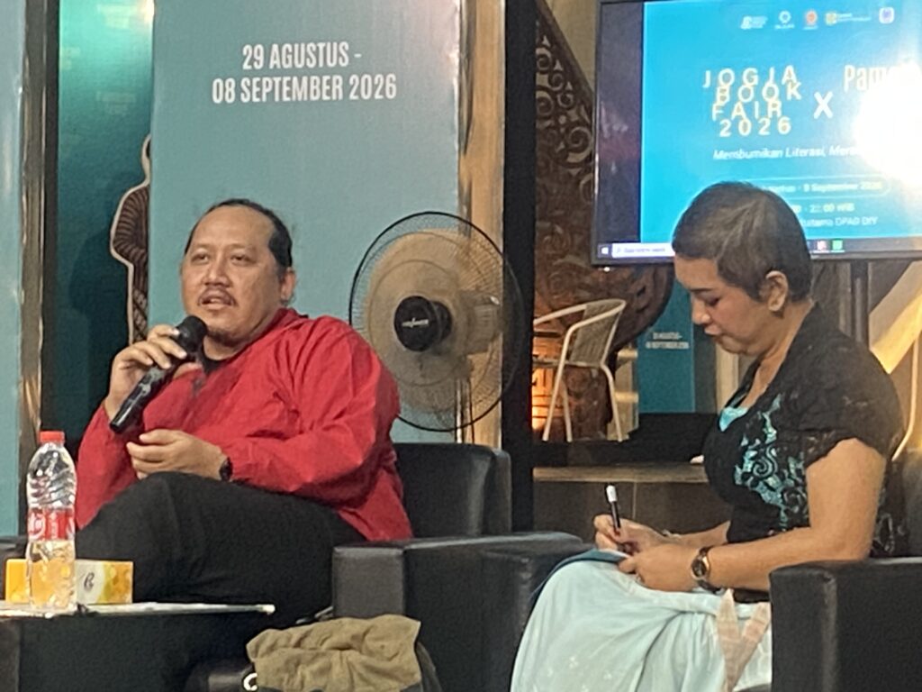 IMG_3116 - Mabur.co Man in a red shirt speaks into a microphone while seated at a panel, with a woman taking notes nearby and event banners in the background.