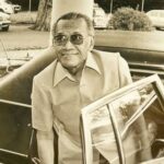Older man with sunglasses exiting a vintage black car in a parking lot, smiling.