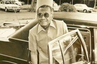 Older man with sunglasses exiting a vintage black car in a parking lot, smiling.