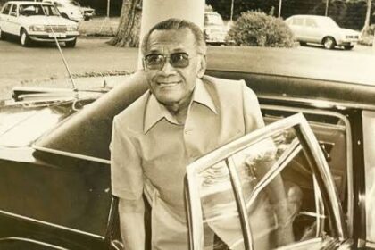Older man with sunglasses exiting a vintage black car in a parking lot, smiling.