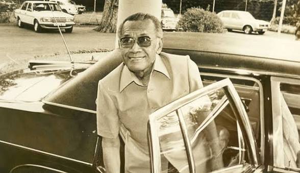 Older man with sunglasses exiting a vintage black car in a parking lot, smiling.