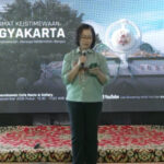 Female presenter on a stage, holding a microphone, in front of a banner that reads 'Maklumat Keistimewaan Yogyakarta' with decorative backdrop and a patterned carpet.